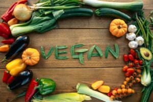 Fresh vegetables arranged on a wooden table spelling the word “VEGAN,” surrounded by colorful produce like peppers, zucchini, tomatoes, garlic, and