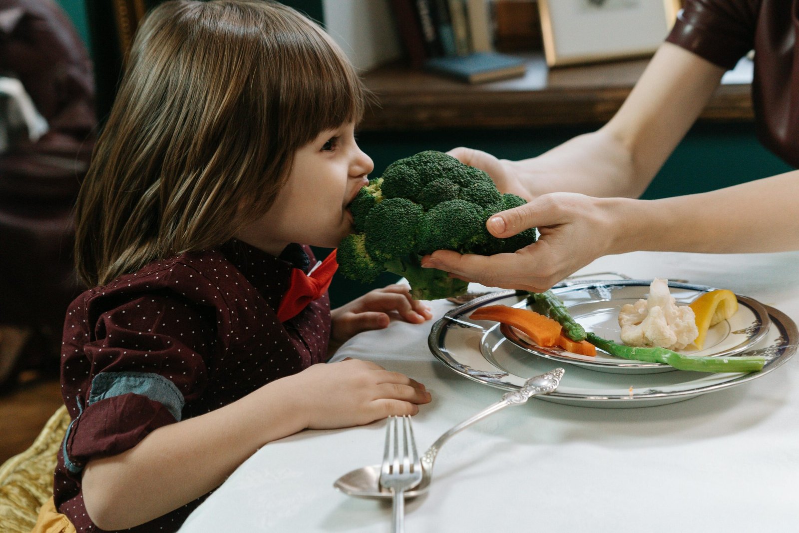 A young child sitting at a dining table while an adult gently helps them eat fresh vegetables, symbolizing the importance of small healthy habits, care, and guidance for long-term personal growth