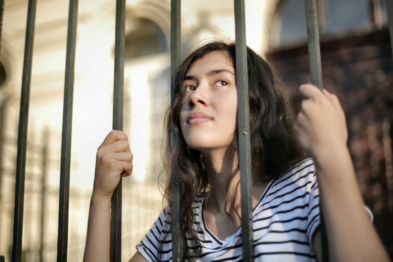 A young woman with long dark hair stands behind a metal fence, lightly holding the bars with both hands and gazing upward, wearing a striped T-shirt with a sunlit building blurred in the background