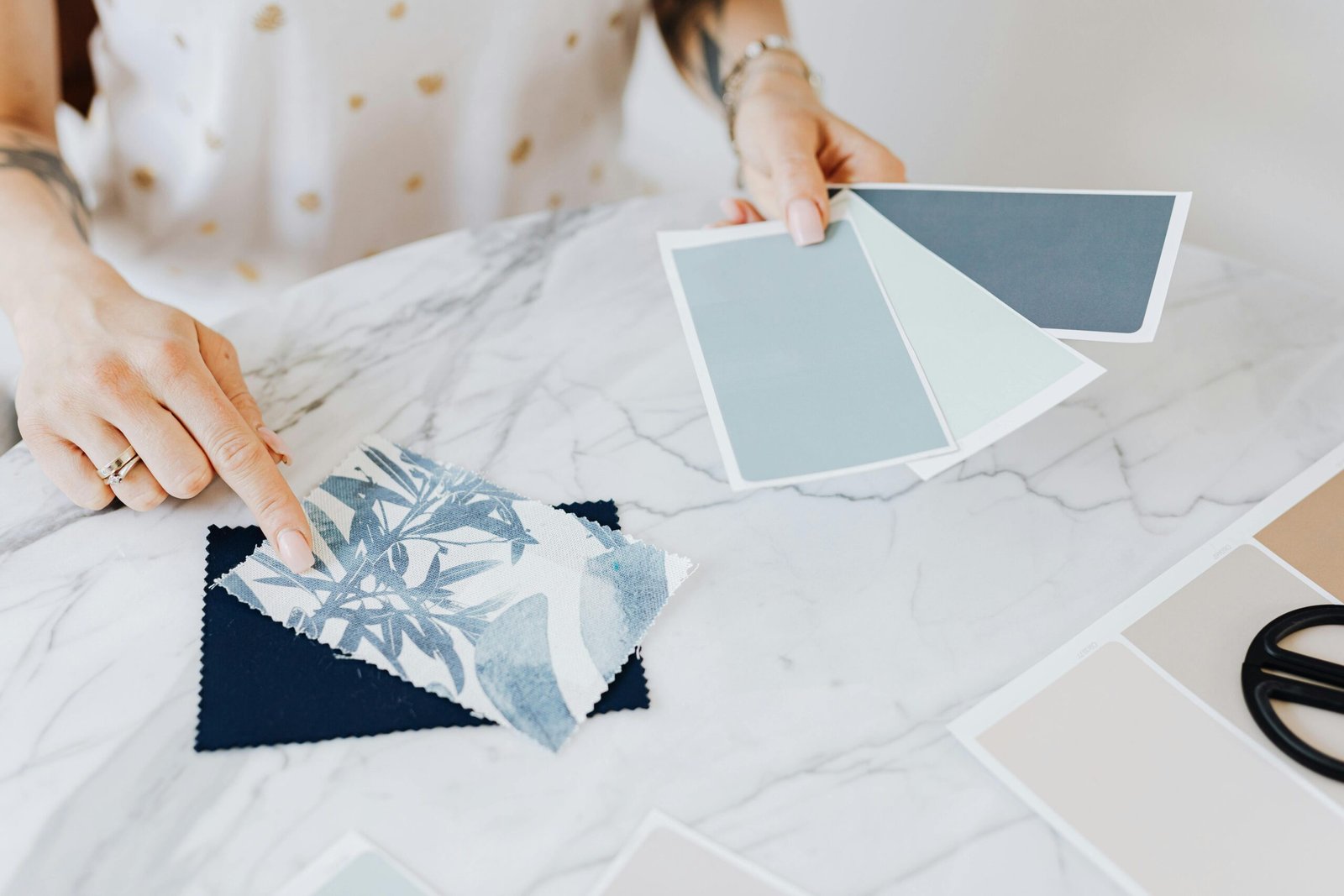 Hands choosing fabric swatches and blue-gray color palette cards on a white marble table, representing design planning and color selection