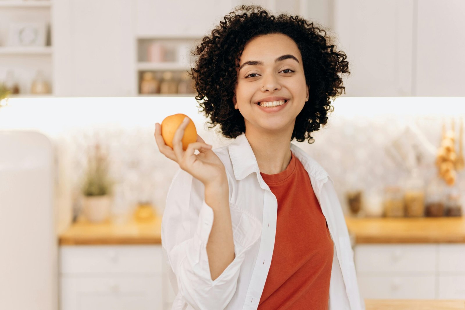 A smiling woman in a bright kitchen holding an orange, representing simple, healthy habits and a stress free lifestyle.