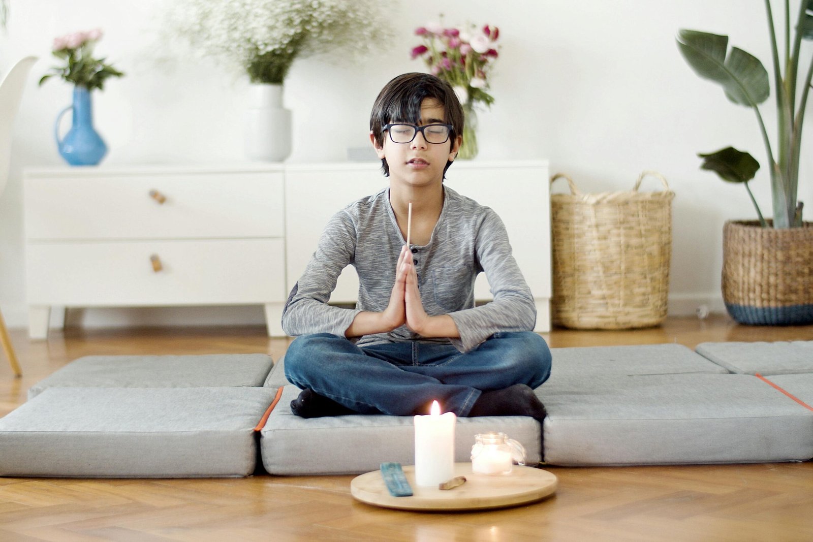 A child wearing glasses sits cross-legged on floor cushions indoors, eyes closed and hands pressed together in a calm meditation pose, with a lit candle and incense on a small tray in front, and plants and flowers in the background