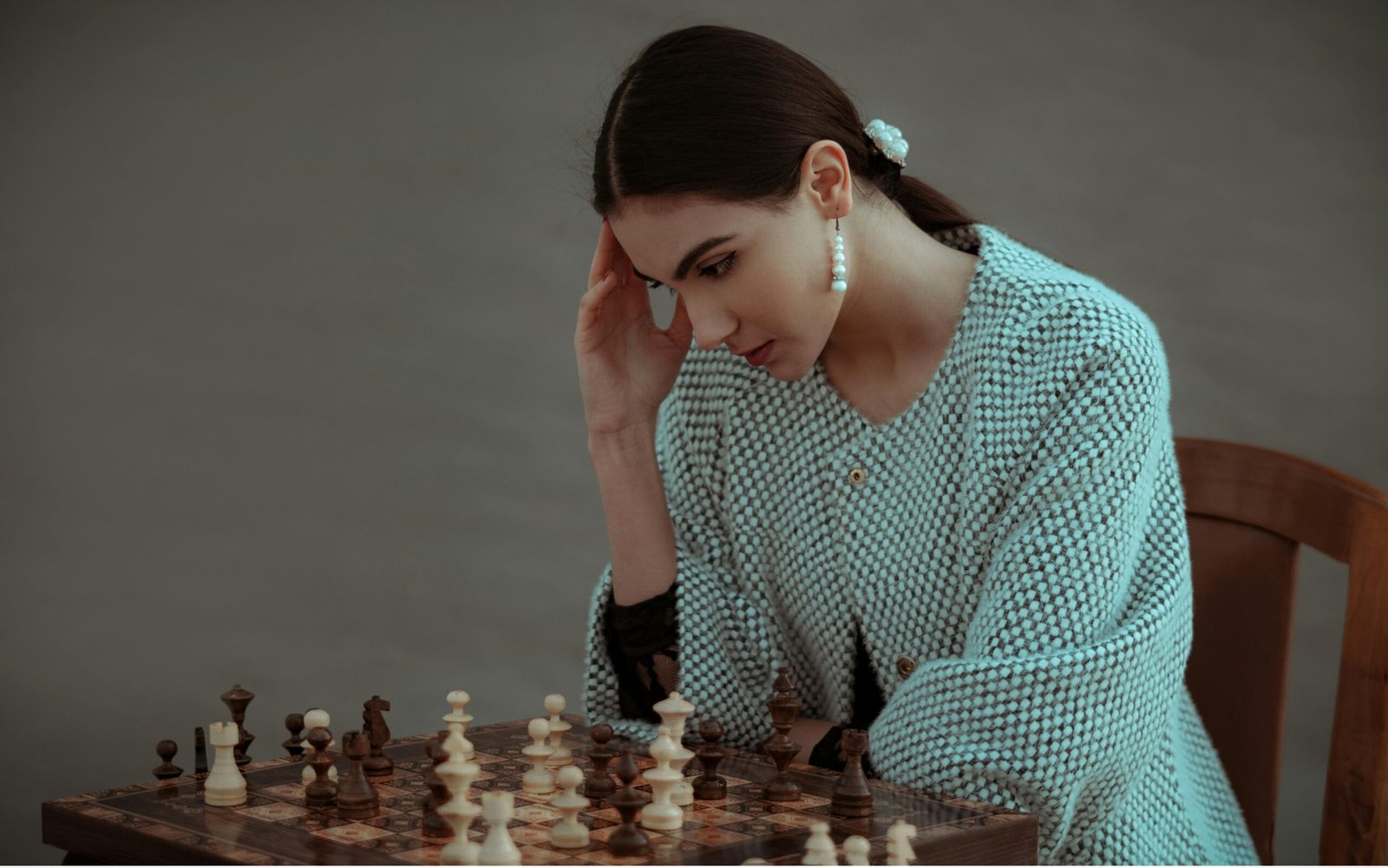 A focused woman sitting at a table, thoughtfully studying a chessboard, resting her hand on her temple while planning her next move in a calm, minimalist setting.